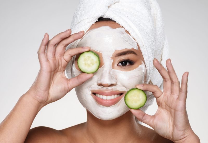 Attractive smiling lady with scrab on face holding cucumber slices isolated Attractive smiling asian lady with scrab on face and towel on head holding cucumber slices isolated over grey